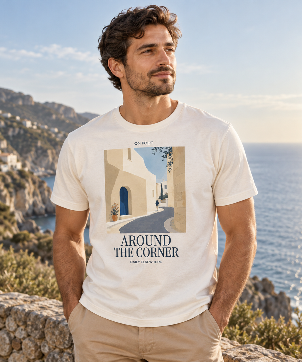 Man wearing an Natural Raw Around The Corner T-shirt with a blue-door village lane print and dark lettering beside the coast.