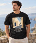 Man wearing a Black Around The Corner T-shirt with a village lane print and light lettering beside the coast.