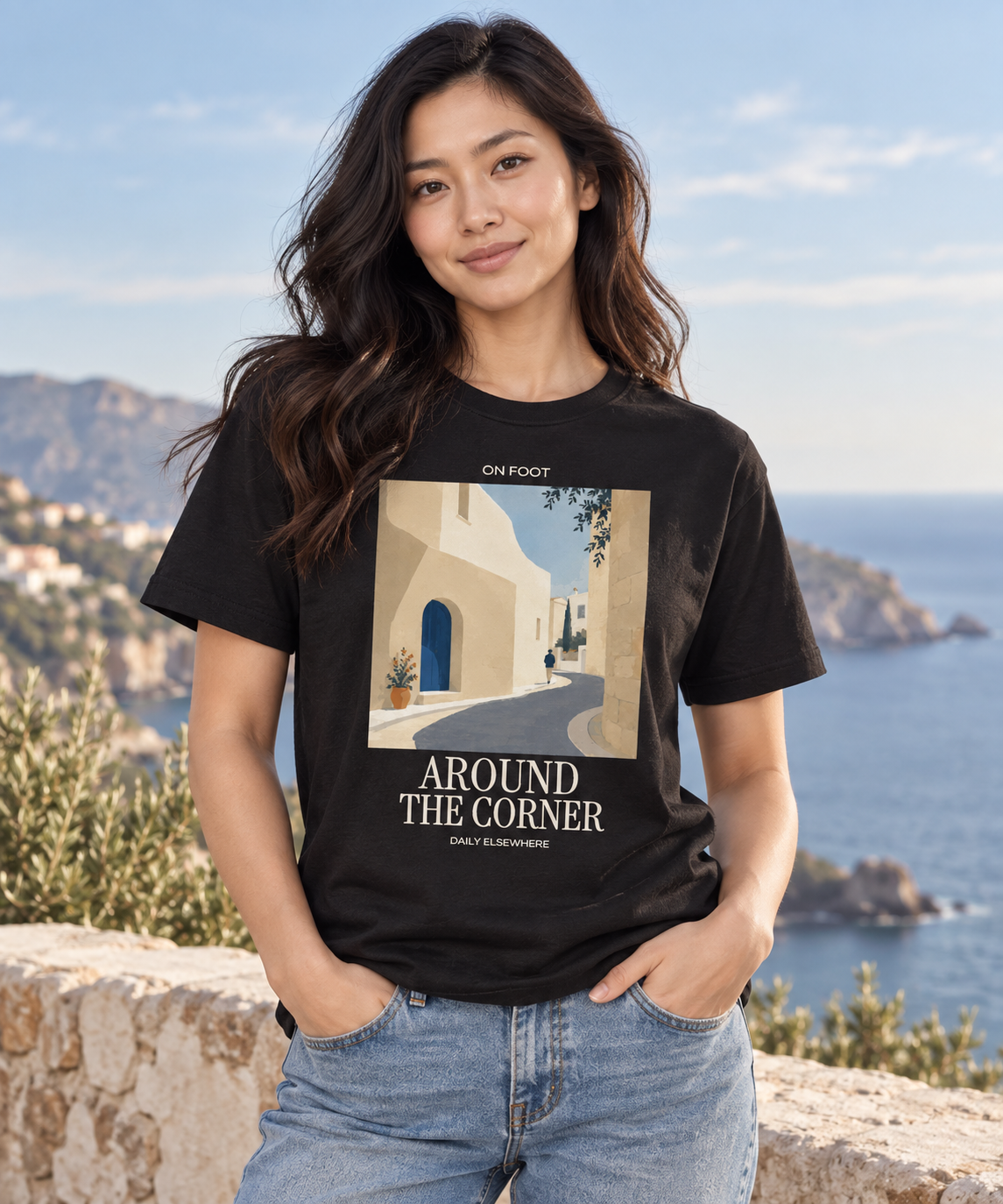 Woman wearing a Black Around The Corner T-shirt with a village lane print and light lettering beside the coast.