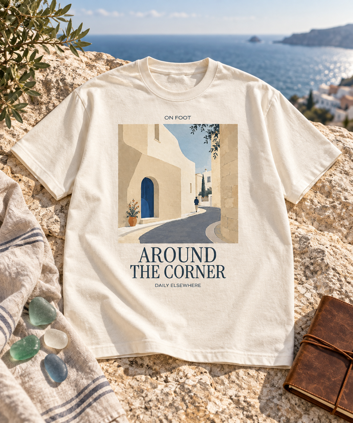 Natural Raw Around The Corner T-shirt with a village lane print and dark lettering laid on coastal stone beside olive branches.