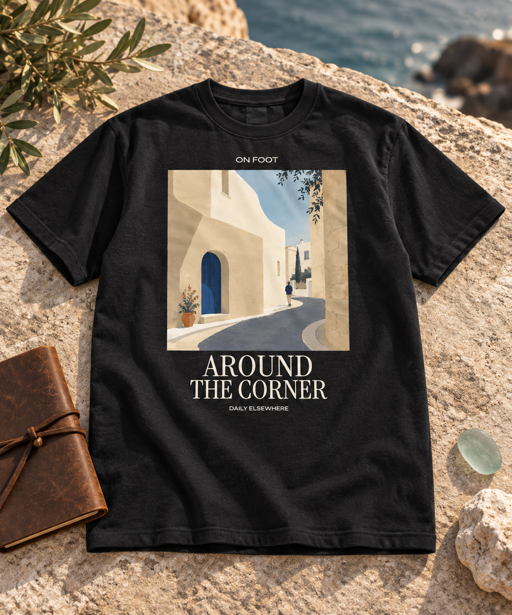 Black Around The Corner T-shirt with a village lane print and light lettering laid on coastal stone beside olive branches.