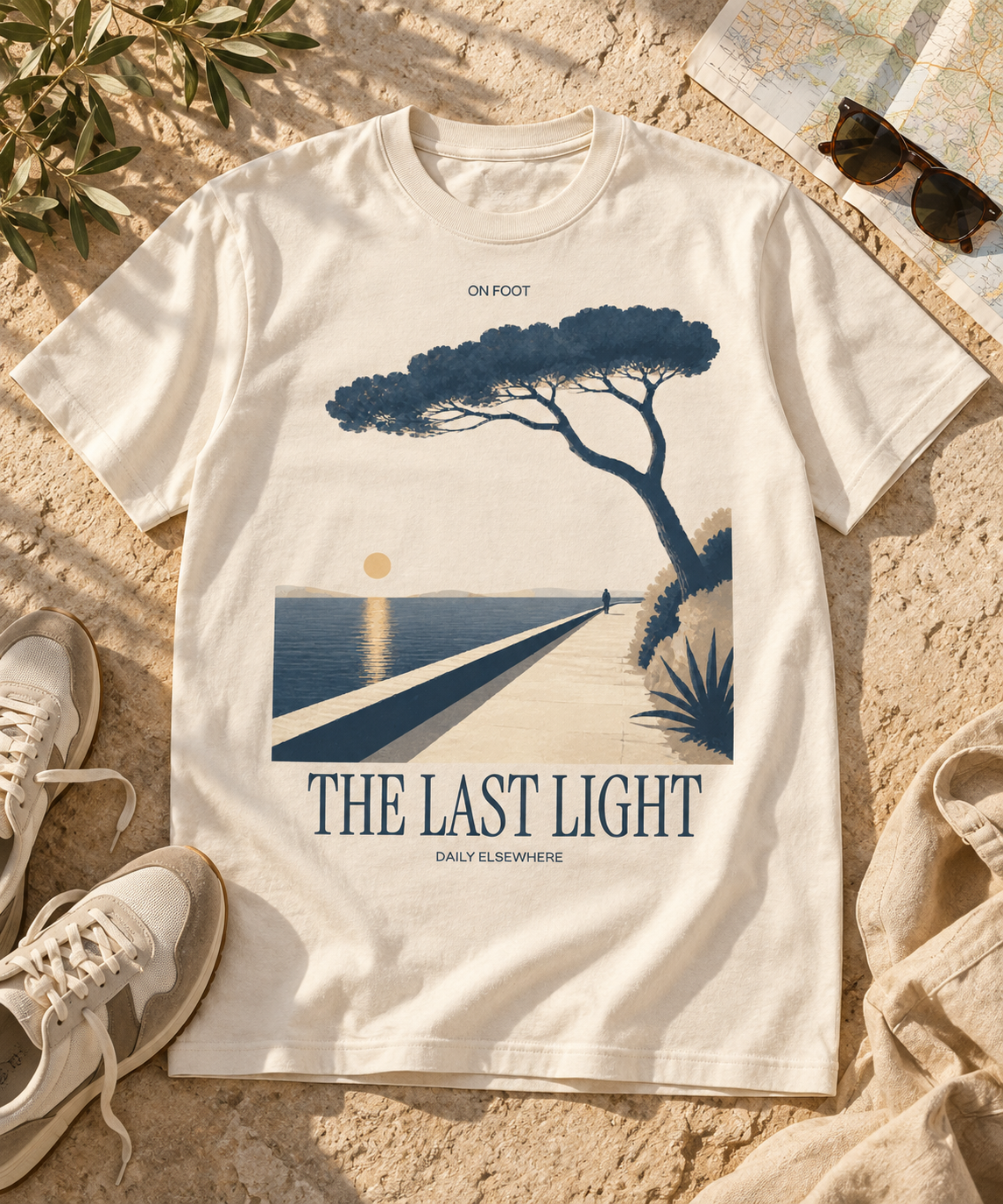 The Last Light Natural Raw T-shirt with dark front artwork, styled on pale stone with travel accessories.
