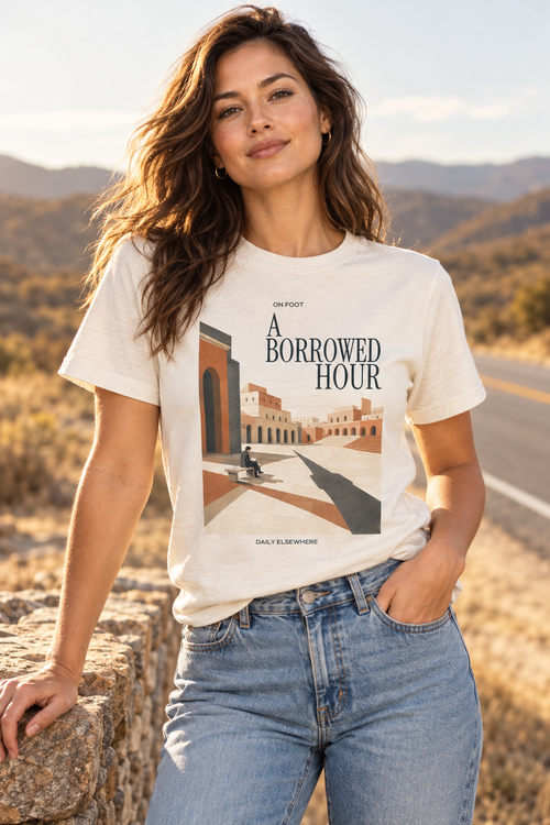 Female model wearing the A Borrowed Hour Natural Raw T-shirt with dark title lettering and a seated reader in a courtyard.
