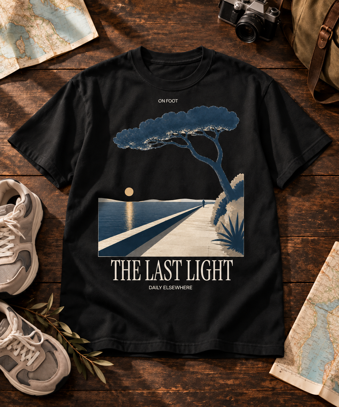 The Last Light black T-shirt with light front artwork, arranged with a camera and map on a wooden table.