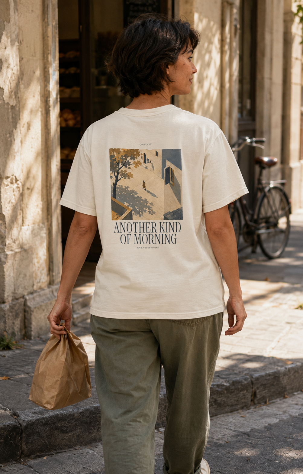 Illustrative lifestyle back view of the Ivory Comfort Colors 1717 T-shirt: Another Kind of Morning artwork during a morning walk outside a neighborhood bakery.